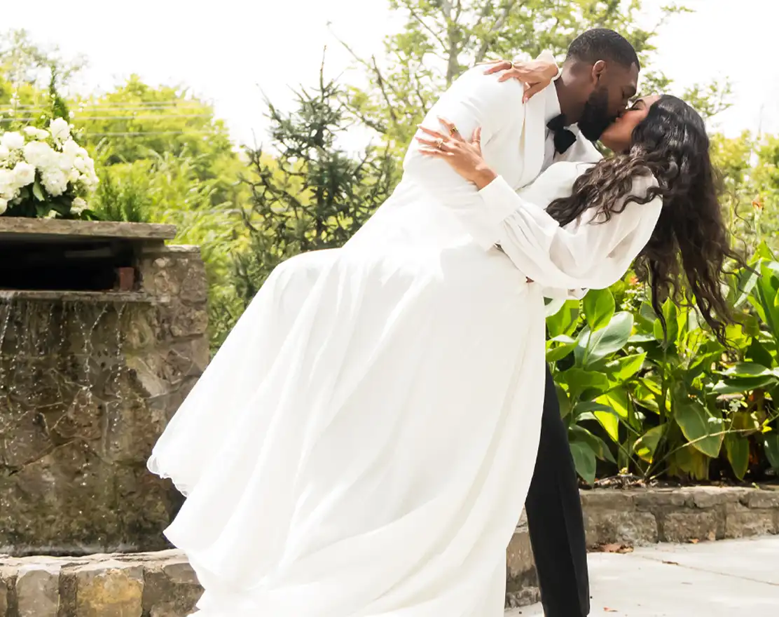 Groom dipping his bride for a kiss beside a stone garden fountain